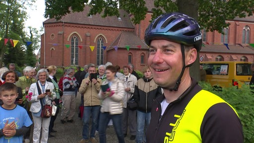 Der Bremer Pastor Pawel Nowak lächelt mit seinem Helm auf dem Kopf in die Kamera. Im Hintergrund stehen viele Menschen vor einer Kirche.