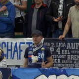 Ein Mann mit Trikot und Schal der Eisbären Bremerhaven steht vor einem Banner und haut mit einem Schlegel auf eine Trommel