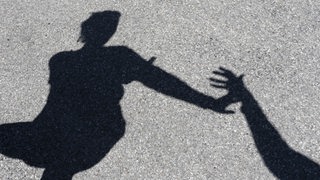 Ein Schattenbild auf dem Asphalt zeigt den Arm eines Mannes, der nach einer Frau greift. 