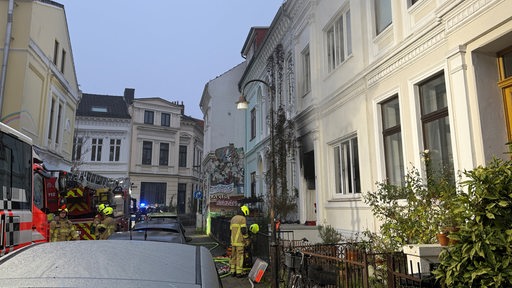 Die Feuerwehr steht in einer schmalen Straße vor einem Haus, dessen Eingang von Rauch schwarz ist