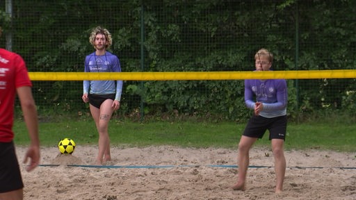 Zwei Männer stehen hinter dem Netz eines beachvolleyballplatzes, während ein Ball auf einem Sandhügel liegt.