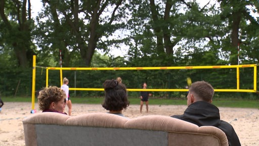 Drei Personen auf einer Couch vor einem Volleyballplatz. 