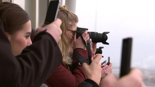 Eine blonde Frau, die ein Foto mit einer Kamera macht, um sie herum stehen mehrere Menschen mit Smartphones in der Hand.