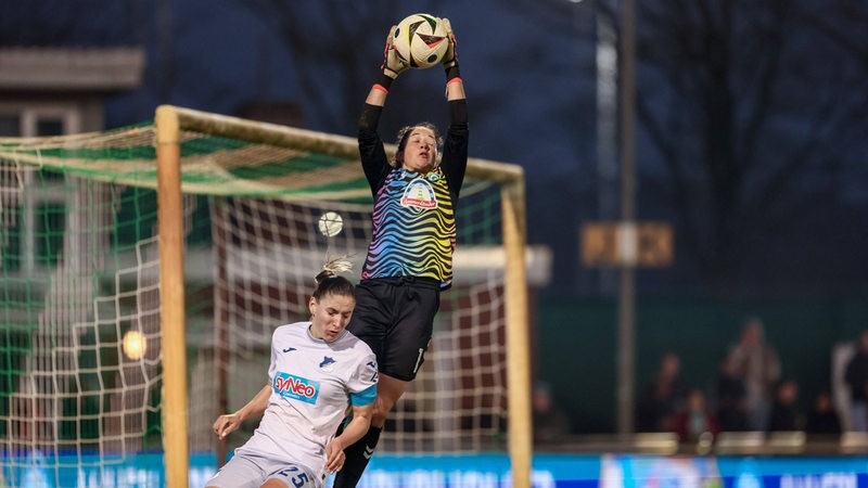 Werders Torhüterin springt hoch in die Luft und schnappt sich den Ball mit hochgereckten Armen und wird dabei von einer Hoffenheimer Spielerin bedrängt.