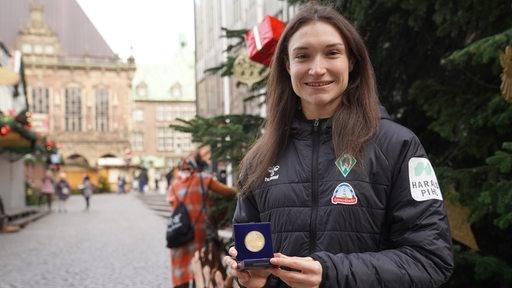 Werder-Stürmerin Sophie Weidauer hält strahlend auf dem Bremer Weihnachtsmarkt ihre Medaille für das "Tor des Monats" der ARD-Sportschau in die Kamera.