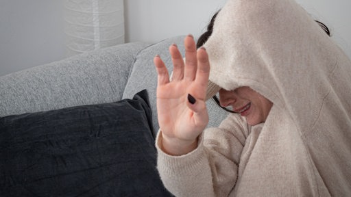 Eine Frau auf dem Sofa hält sich ängstlich schützend die Arme vor ihr Gesicht.