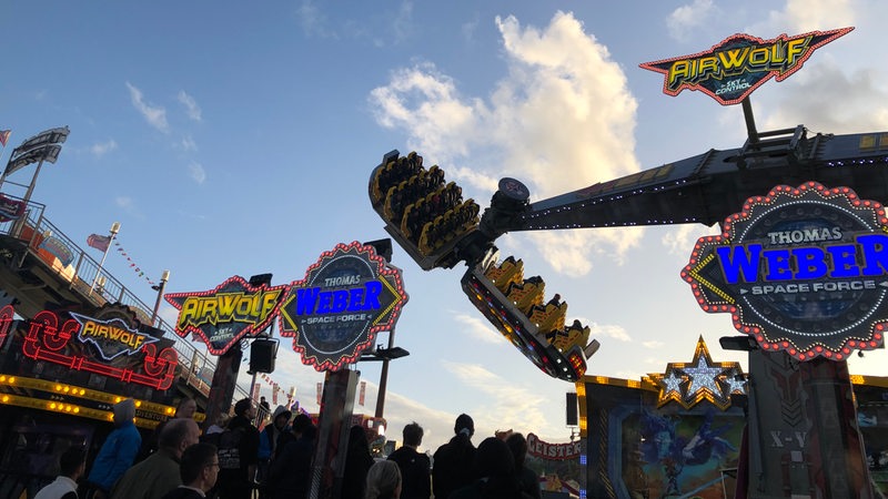 Das Fahrgeschäft "Airwolf" ist erleuchtet vor dem dunkler werdenen Himmel zu erkennen. Im Vordergrund sind Menschen als Schattenrisse zu erkennen.