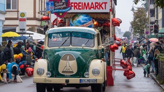 Ein geschmückter Wagen mit der Aufschrift "Freimarkt" führt den Bremer Freimarktsumzug an, Menschen jubeln ihm zu.