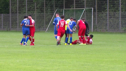 Auf einem Fußballspielfeld sind zwei Mannschaften zu sehen. Ein Junge liegt auf dem Boden und seine Mitspieler stehen um ihn herum.