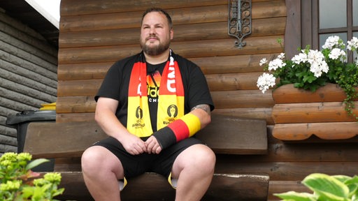 Ein Fußballfan sitzt mit Deutschland-Schal vor einer Holzhütte.