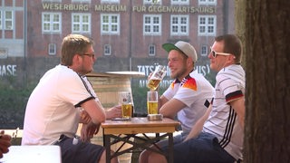 Drei Personen sitzen im Trikot der deutschen Fußball-Nationalmannschaft im Biergarten an der Schlachte.