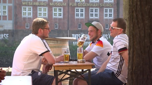 Drei Personen sitzen im Trikot der deutschen Fußball-Nationalmannschaft im Biergarten an der Schlachte.