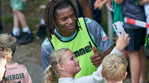 Werder-Profi Skelly Alvero posiert auf dem Weg zum Training freundlich für das Selfie eines Mädchens.