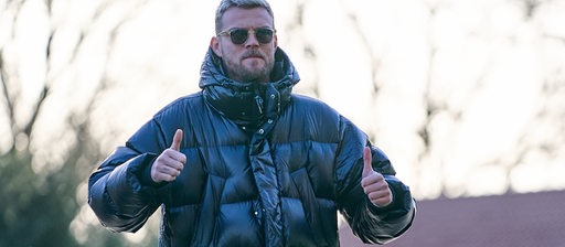 Werder-Stürmer Marvin Ducksch reckt auf dem Weg zum Weser-Stadion in einer dunklen Daunenjacke und mit Sonnenbrille beide Daumen hoch.