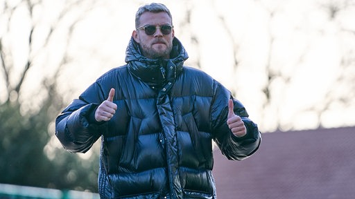 Werder-Stürmer Marvin Ducksch reckt auf dem Weg zum Weser-Stadion in einer dunklen Daunenjacke und mit Sonnenbrille beide Daumen hoch.