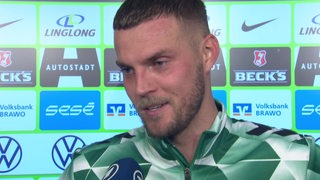 Werder-Profi Marvin Ducksch lächelt beim Interview nach dem Sieg in Wolfsburg vor einer Werbewand.