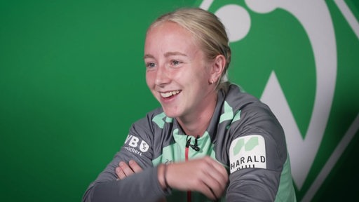 Die Werderspielerin Caroline Siems sitzt lächelnd vor einer grünen Wand mit Werder-Raute.