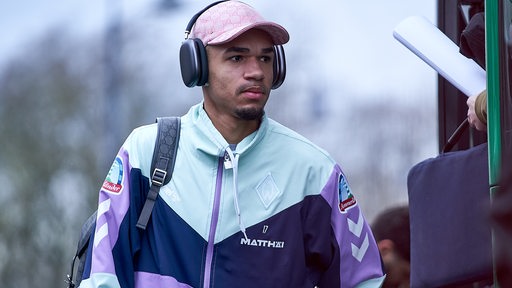 Werder-Spieler Justin Njinmah lässig in rosa-lilafarbener Freizeitkleidung mit Kappe und Kopfhörer auf dem Weg zum Weser-Stadion.