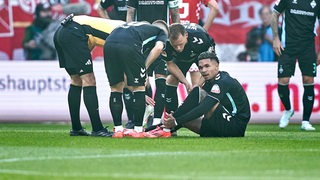 Werder-Stürmer Justin Njinmah sitzt auf dem Rasen während des Spiels in Mainz und hält sich den Knöchel, andere Mitspieler und der Schiedsrichter erkundigen sich, was los ist.