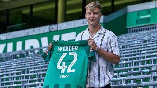 Werders Neuzugang Keke Topp hält in der leeren Ostkurve im Weser-Stadion sein Trikot mit der Rückennummer 42 hoch.