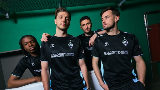 Vier Werder-Spieler posieren im neuen Werder-Trikot, das hauptsächlich schwarz ist.