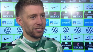 Werder-Profi Mitchell Weiser lächelt beim Interview nach dem Sieg in Wolfsburg vor einer Werbewand.