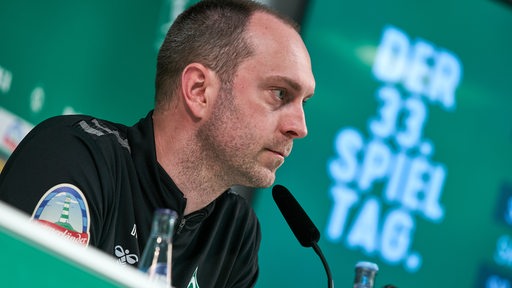 Werder-Trainer Ole Werner sitzt auf dem Podium einer Pressekonferenz, hinter ihm ein Bildschirm mit der Schrift "Der 33. Spieltag".