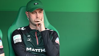 Werder-Trainer Ole Werner sitzt nachdenklich mit auf der Trainerbank, das Gesicht auf seine Hand gestützt.