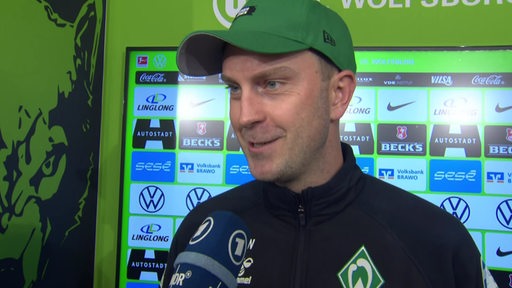 Werder-Trainer Ole Werner lächelt beim Interview nach dem Sieg in Wolfsburg vor einer Werbewand.