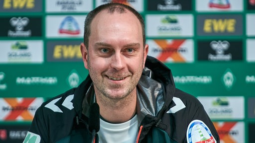 Werder-Trainer Ole Werner sitzt lächelnd bei einer Pressekonferenz.