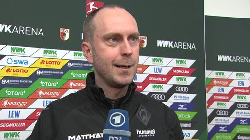Werder-Trainer Ole Werner steht nach dem Spiel lächelnd vor einer Werbewand beim Interview.