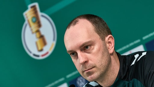 Werders Trainer Ole Werner sitzt bei einer PK. Im Hintergrund ist das Logo des DFB-Pokals zu sehen.