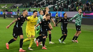 Werder-Fußballer laufen nach dem Sieg in Wolfsburg aufgereiht nebeneinander in Richtung Fankurve und feiern mit ihnen.