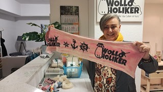 Eine Frau hält einen Schal mit der Aufschrift ""Wolloholiker" in die Kamera.