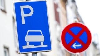 Ein Verkehrszeichen erlaubt das aufgesetzte Parken auf dem Gehweg, daneben steht ein Schild, das ein absolutes Halteverbot anzeigt. 