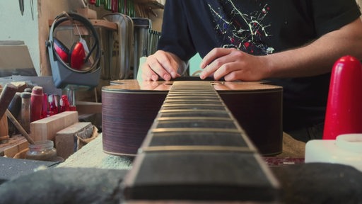 In einer Werkstatt klebt eine Person etwas auf den Körper einer unfertigen Gitarre