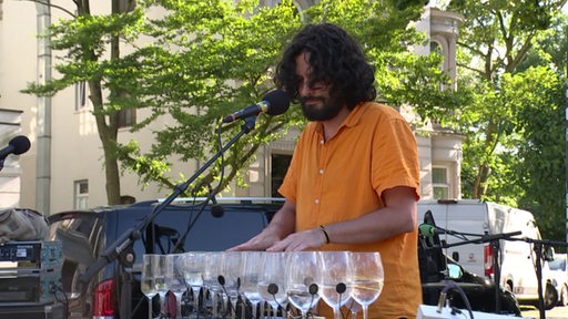 Ein Musiker spielt auf einer selbstgebauten Glasharfe bestehend aus vielen mit Wasser gefüllten Weingläsern.