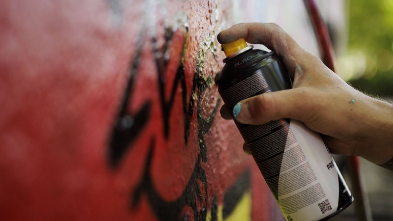 Ein Mann sprueht mit einer Spraydose ein Graffiti an eine Wand. 