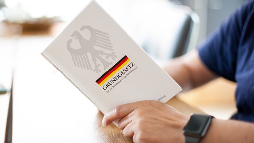 Eine Person hält das Grundgesetz, GG, der Bundesrepublik Deutschland, in den Händen und liest darin an einem Tisch.