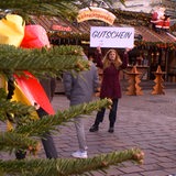 Eine junge Frau hält ein Schild mit dem Wort "Gutschein" in die Höhe. Im Vordergrund sind Zweige eines Tannenbaums zu sehen