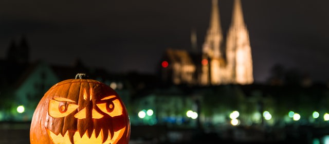 Ein Halloween-Kürbis liegt bei Dunkelheit auf einer Mauer. Im Hintergrund sieht man eine Kathedrale.