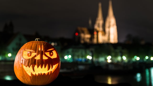 Ein Halloween-Kürbis liegt bei Dunkelheit auf einer Mauer. Im Hintergrund sieht man eine Kathedrale.