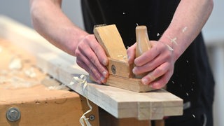 Ein Hobel fährt über ein Holzbrett. Späne fliegen.