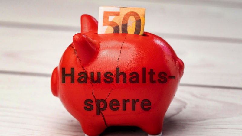 Ein rotes Sparschwein mit Rissen und der Aufschrift "Haushaltssperre" steht auf einem Tisch