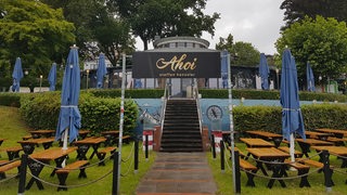 Bänke, Tische und Regenschirme stehen vor einem Restaurant mit dem Namen Ahoi auf grünem Rasen.