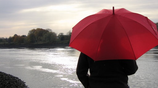 Bei trübem Herbstwetter und Regen schaut eine junge Frau mit rotem Regenschirm über die Weser bei Bremen.