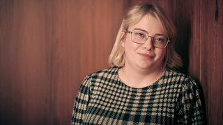 Eine junge Frau mit blonden Haaren lehnt an einer holzvertäfelten Wand und schaut in die Kamera.