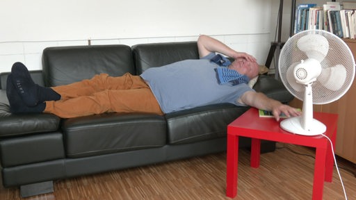 Eine Person liegt neben einem Ventilator auf einem Sofa und hält sich gequält eine Hand ins Gesicht.