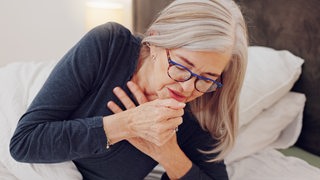 Eine Frau liegt im Bett und beugt sich hustend zur Seite.