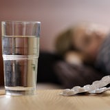 Eine junge Frau liegt krank im Bett, auf dem Nachttisch steht ein Glas Wasser und daneben Schmerztabletten.